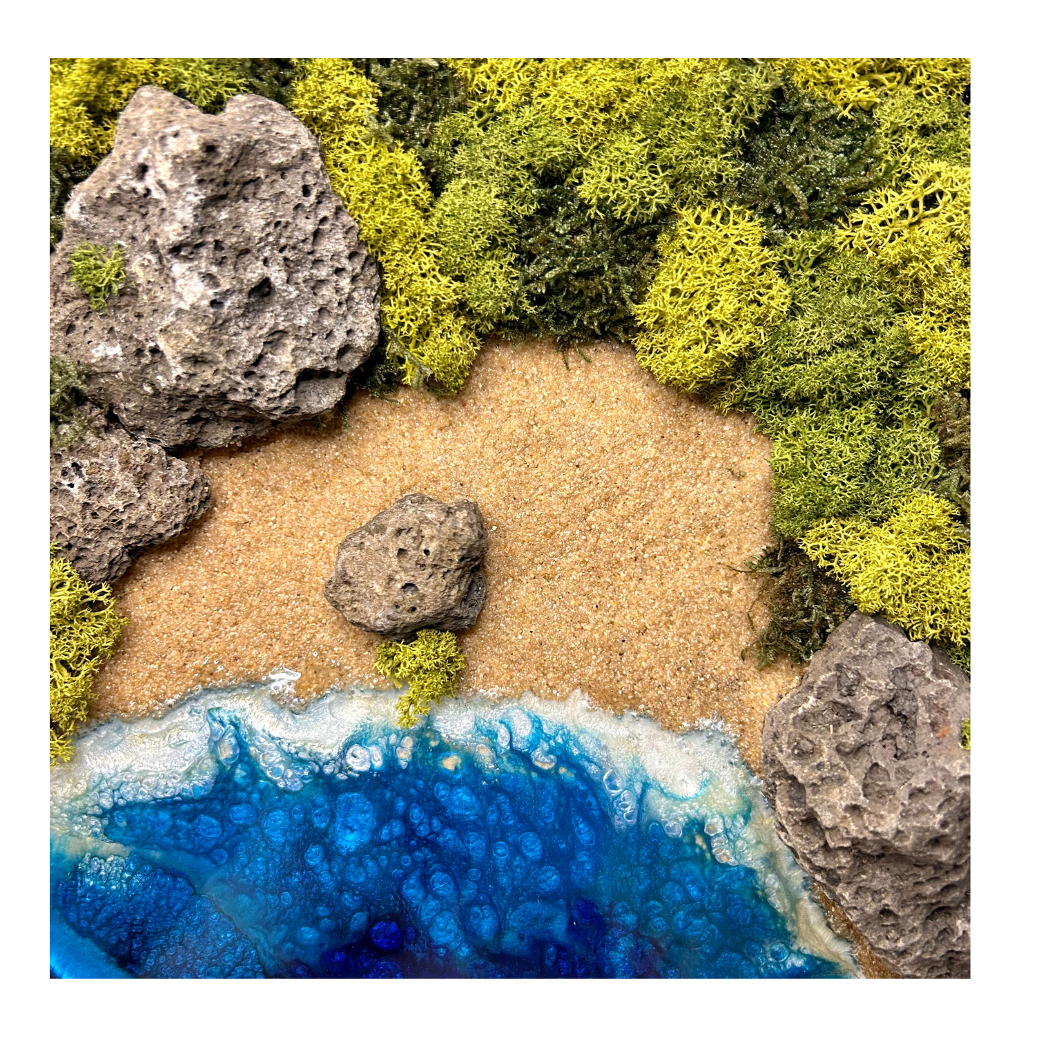 Detailansicht: Moos- und Meerbild "Versteckte Bucht" im schwarzen runden Holzrahmen mit Sandstrand und einem Meer aus blauem Epoxidharz. In das Bild sind Lavasteine eingebaut und mit viel Liebe zum Detail feinste Moosspitzen aus konserviertem Moos eingebaut, die teilweise aussehen wie Palmwedel.