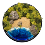 Moos- und Meerbild "Versteckte Bucht" im schwarzen runden Holzrahmen mit Sandstrand und einem Meer aus blauem Epoxidharz. In das Bild sind Lavasteine eingebaut und mit viel Liebe zum Detail feinste Moosspitzen aus konserviertem Moos eingebaut, die teilweise aussehen wie Palmwedel. 