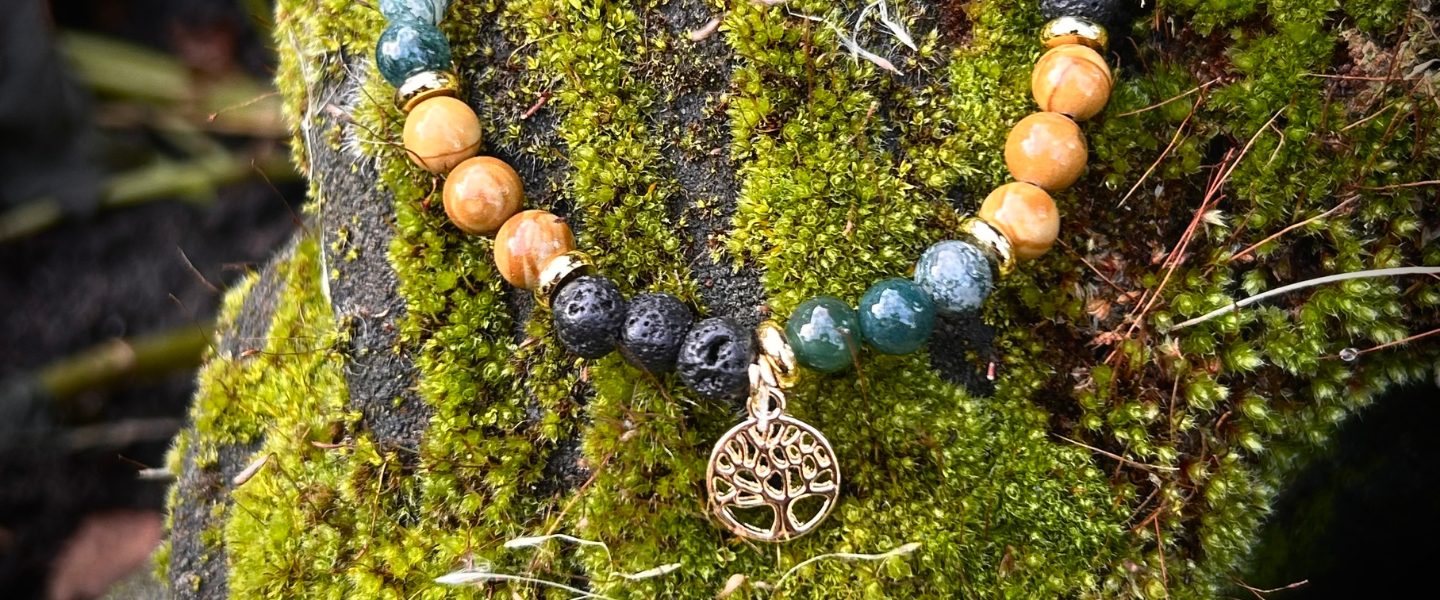 Banner für Moos und Meer Kategorie Schmuck aus Natursteinperlen. HIer siehst du ein Armband aus Moosachat, versteinertem Holz und Lava, mit goldenen Perlen und einem goldenen Anhänger mit dem Baum des Lebens. Das Armband liegt auf einem bemoosten Stein. 