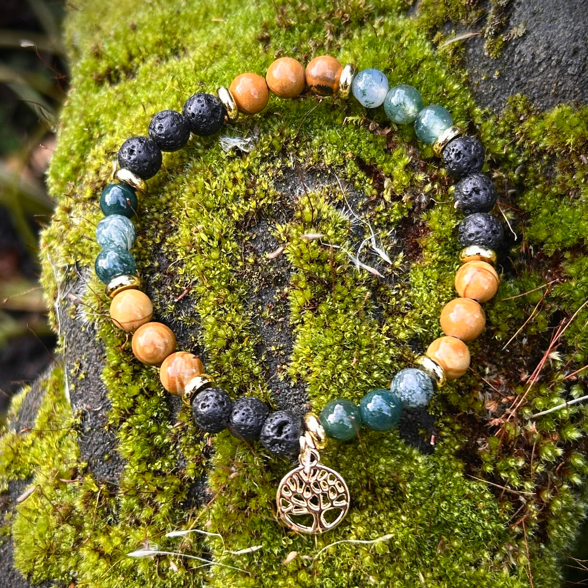 Armband mit versteinertem Holz, Moosachat, Lava und vergoldetem Lebensbaum-Anhänger auf moosbedecktem Stein