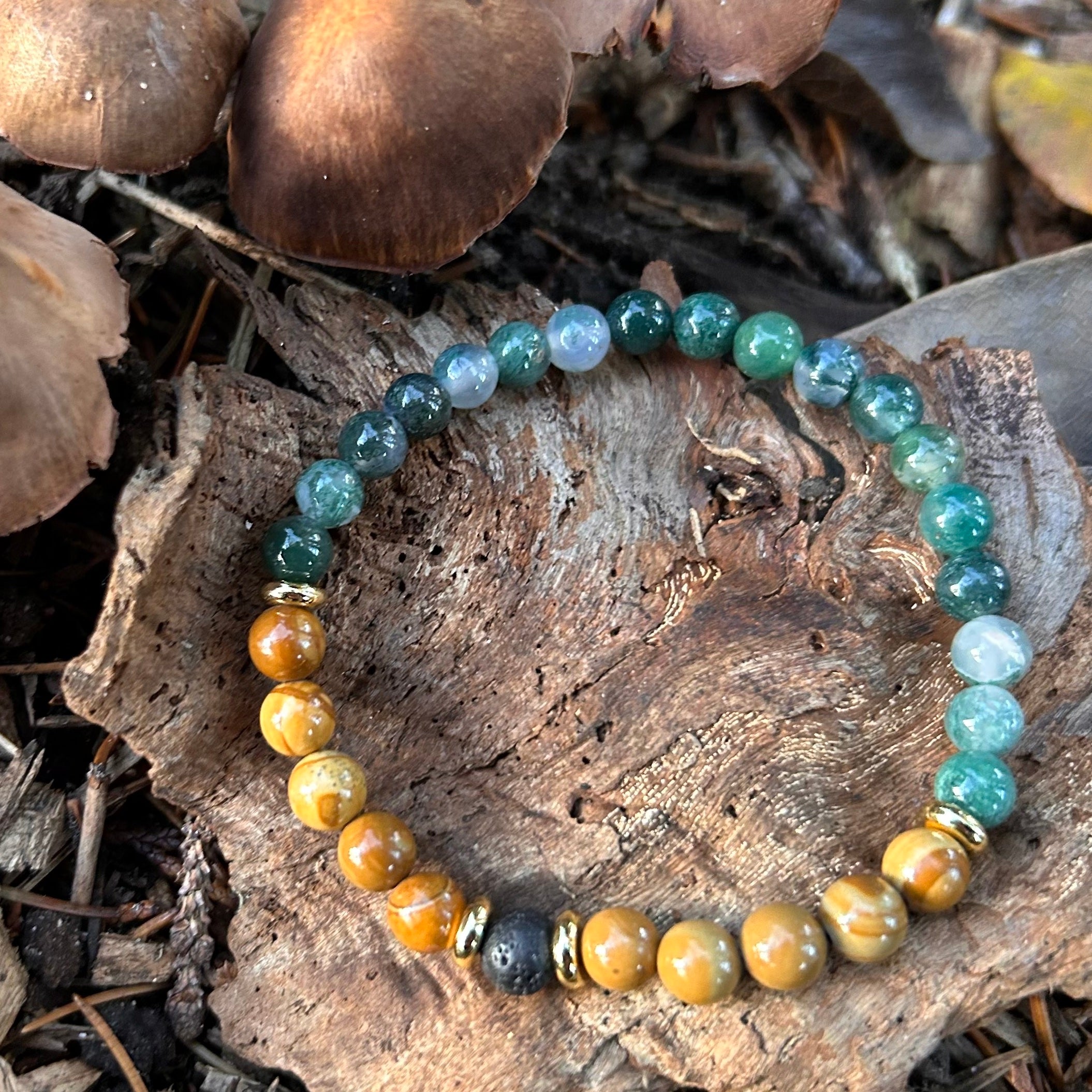 Handgemachtes Armband aus Moosachat, versteinertem Holz und Lava, mit vergoldeten Elementen – fotografiert auf Rinde und Waldboden mit Pilzen