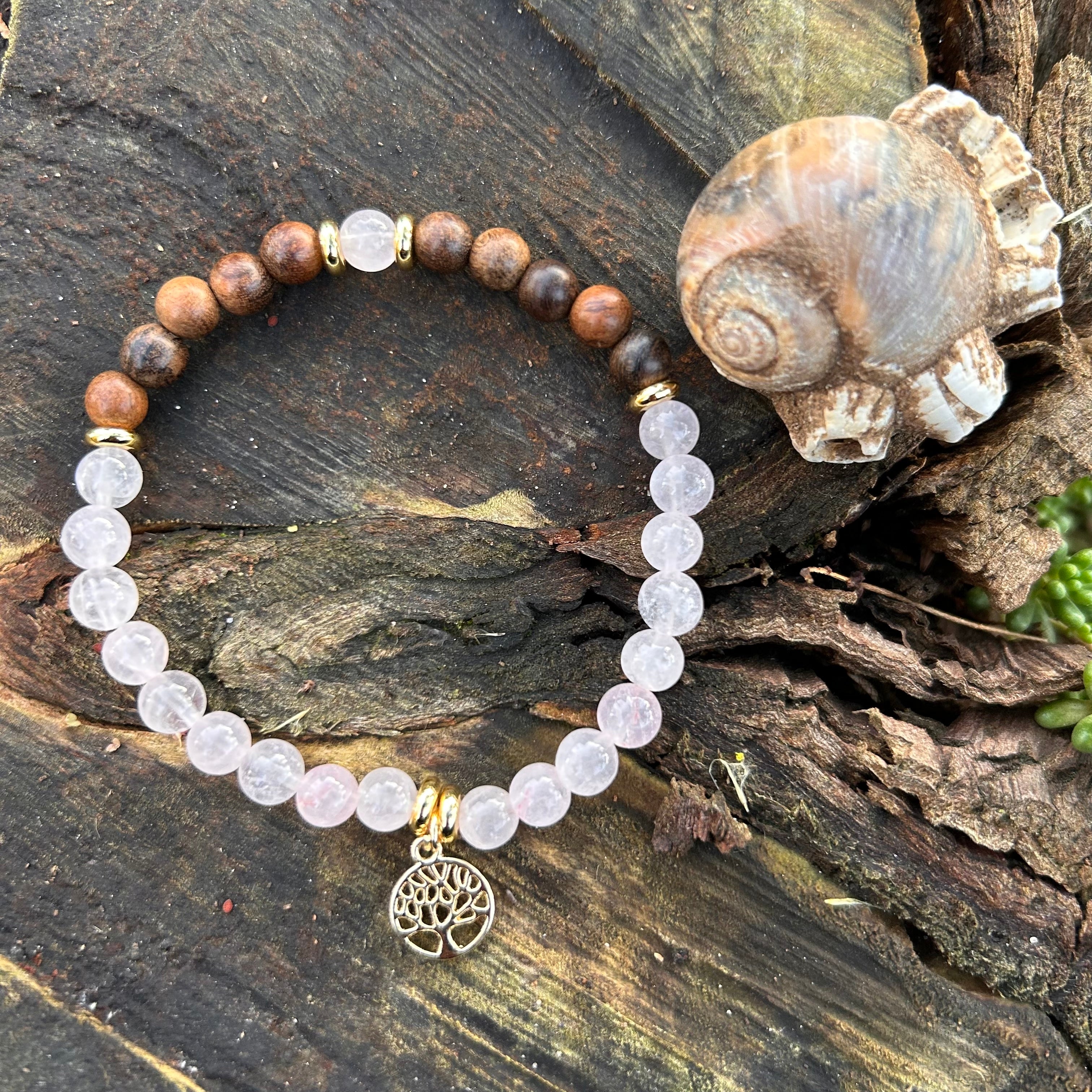 Zartes Armband aus Rosenquarz und Sandelholz mit vergoldetem Lebensbaum-Anhänger auf dunkler Holzrinde, daneben fossile Muschel