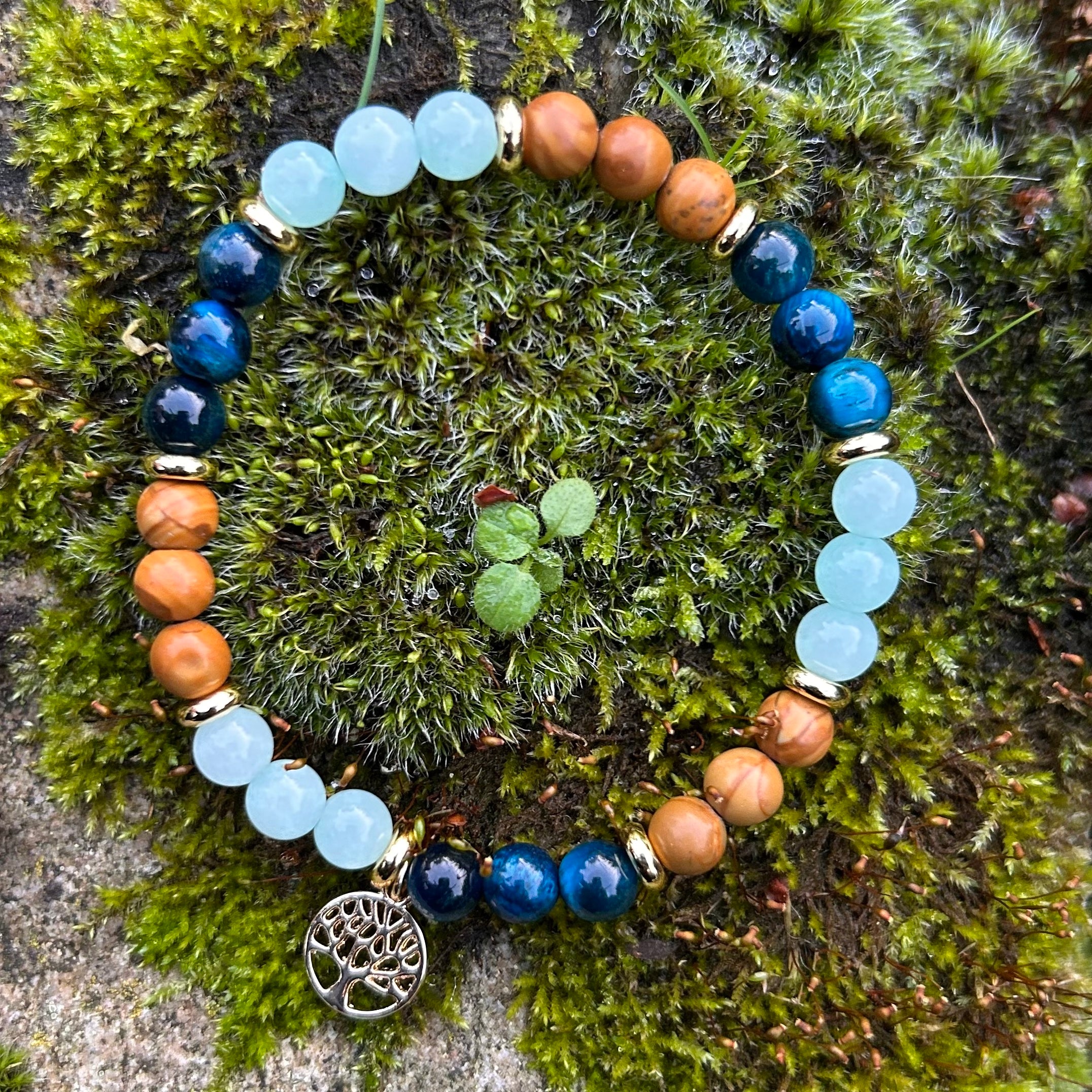 Handgemachtes Edelsteinarmband mit versteinertem Holz, blauem Tigerauge, Amazonit und vergoldetem Lebensbaum-Anhänger auf Holzuntergrund