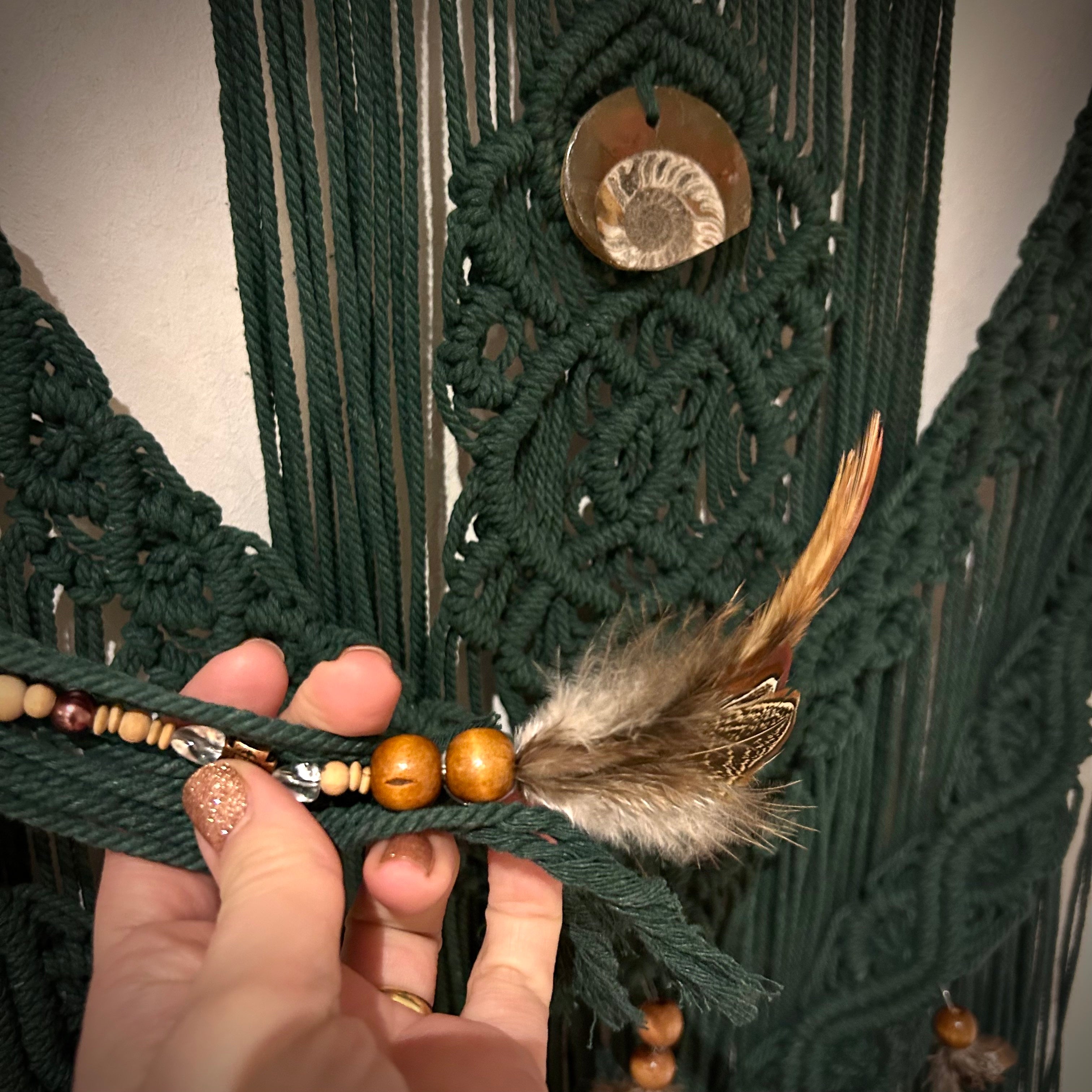 Detailaufnahme: Das dunkelgrüne Makramee im Natur- und Bohostil hängt an einer weißen Wand. Das Makramee wurde an wildes Holz geknüpft und verziert mit Perlen, Federn und einem echten Fossil (Ammonit). 