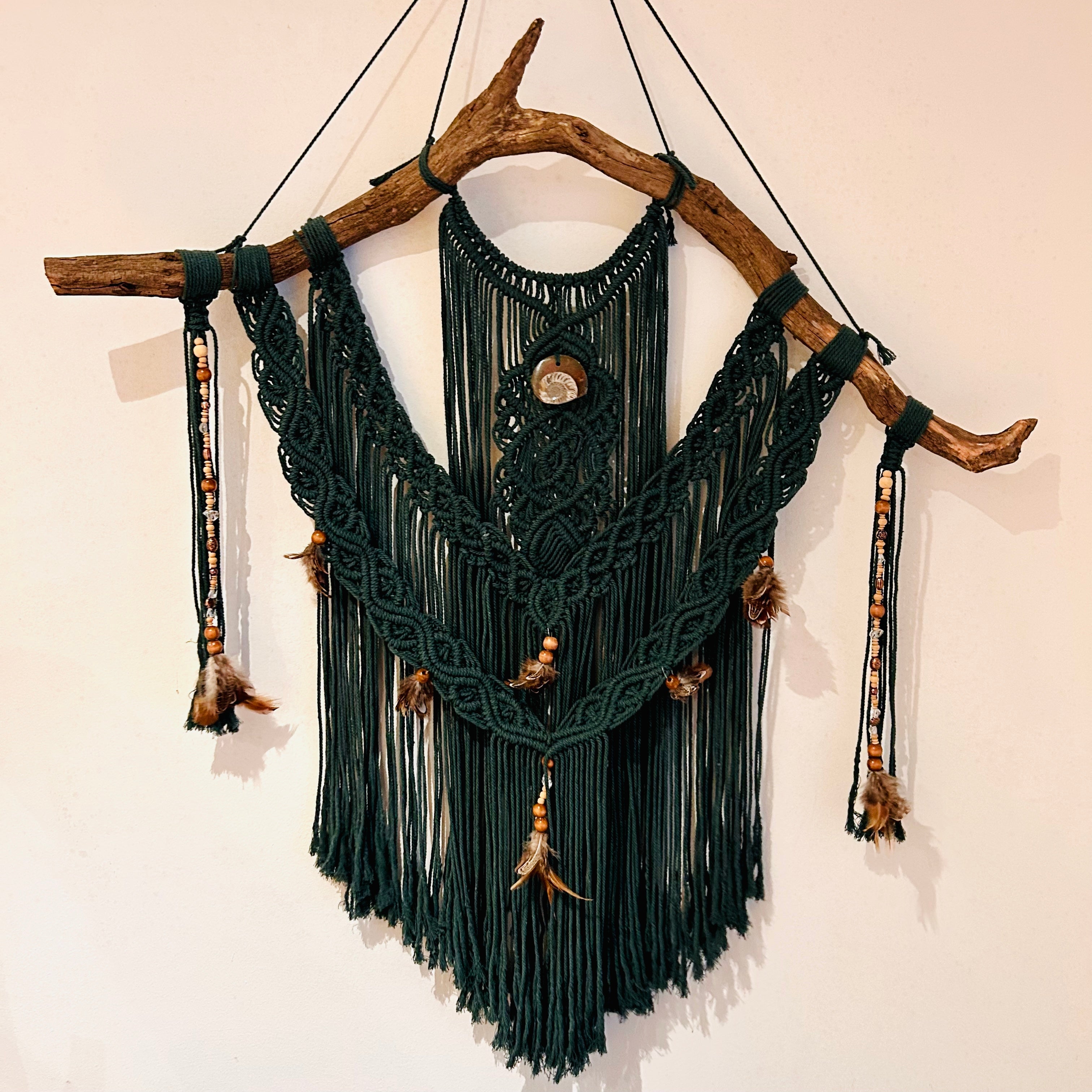 Das dunkelgrüne Makramee im Natur- und Bohostil hängt an einer weißen Wand. Das Makramee wurde an wildes Holz geknüpft und verziert mit Perlen, Federn und einem echten Fossil (Ammonit). 