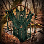 Ein dunkelgrünes Makramee an einem Naturholz hängt in einem Wald. Das Makramee ist aus Baumwolle, verziert mit Perlen, Federn und einem echten Fossil (Ammonit). 