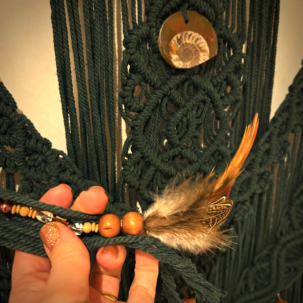 Makramee in Dunkelgrün. Handgeknüpft und mit einem Fossil sowie Perlen und Federn verziert. 