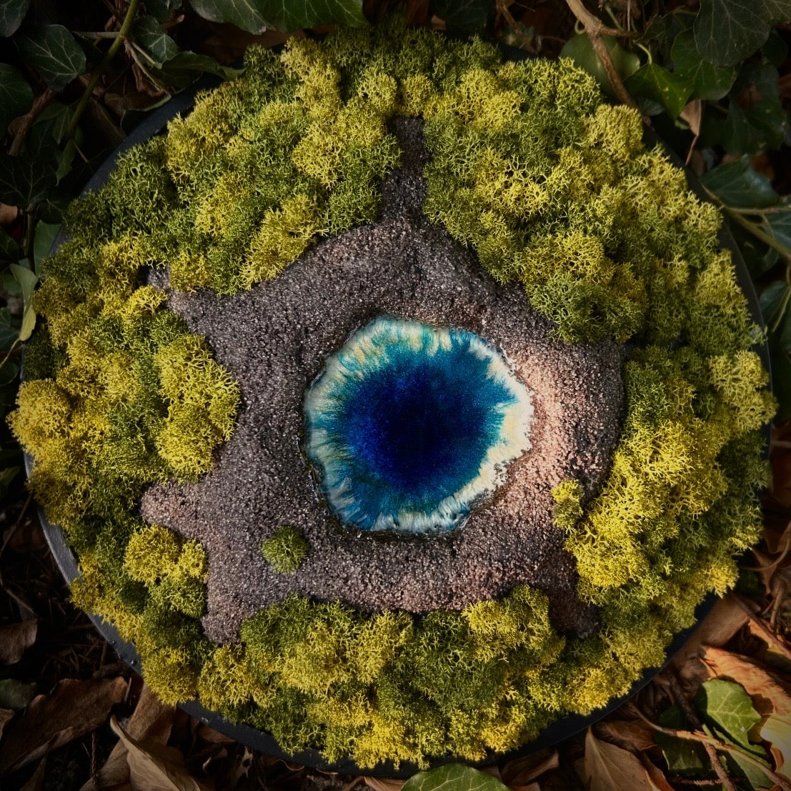 Moosbild Vulkan. Ein Moosbild mit einem von Hand modellierten Vulkan in der Mitte, der sich dimensional aus dem Bilderrahmen heraus bewegt. In der Mitte des Vulkans befindet sich ein Vulkansee aus Resin. 