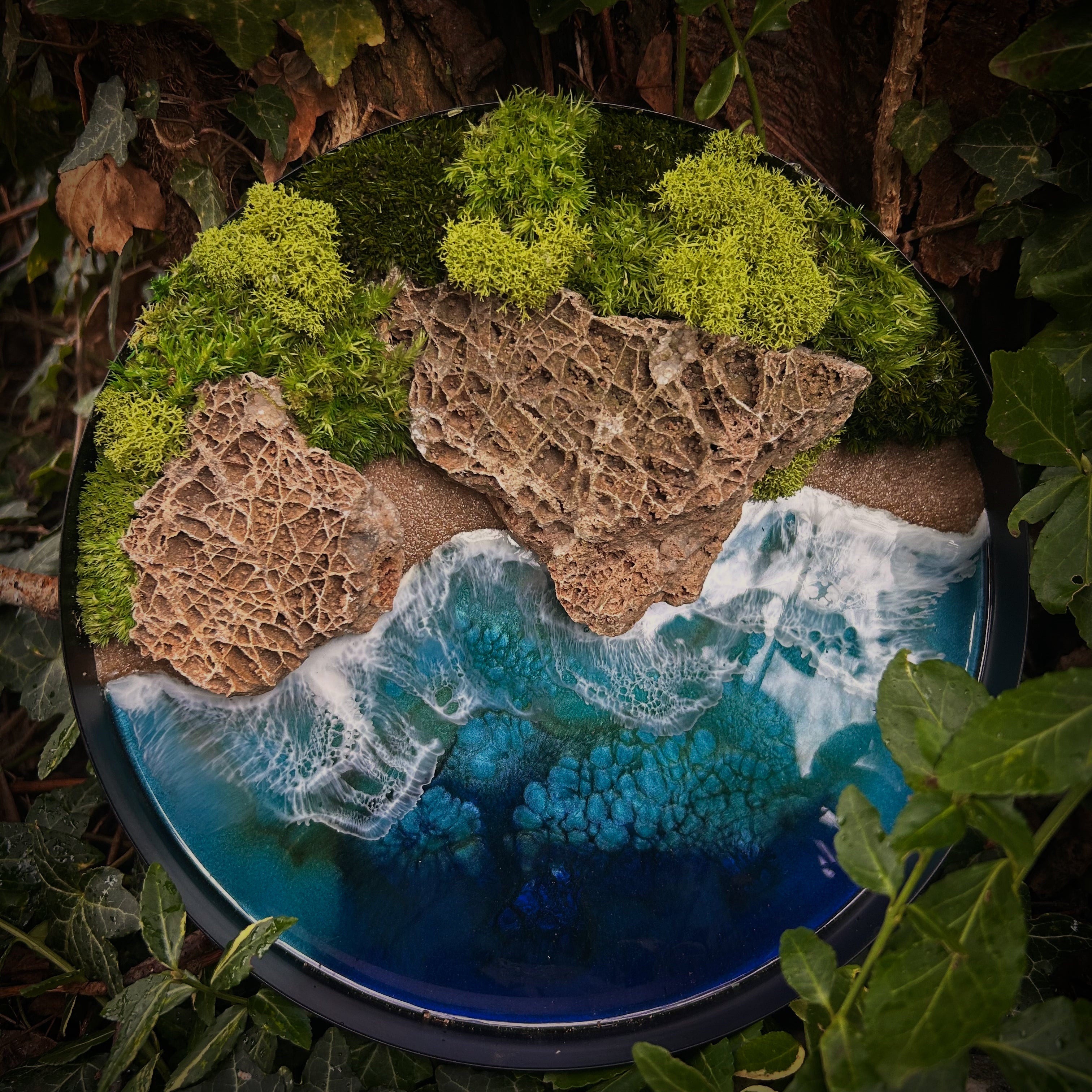 Kleines rundes Moosbild im schwarzen Metallrahmen, das eine maritime Landschaft zeigt. Die Natursteine sind verwittert und bilden ein bizarres Muster. Das Meer aus Resin schlägt am Rand Wellen. Im Hintergrund ist ein Wald aus verschiedenen Moosen. 