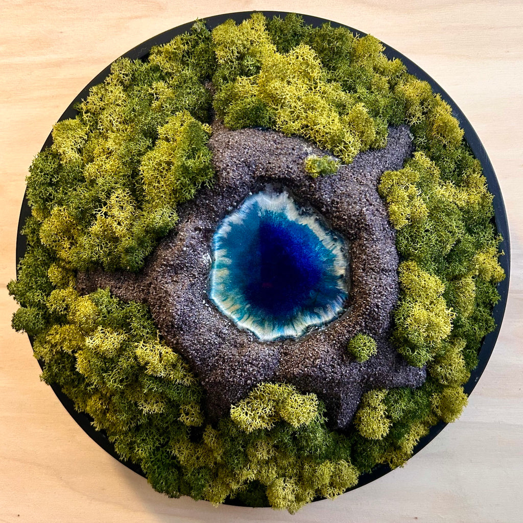 Moosbild "Vulkansee" in einem schwarzen, runden Rahmen. Hier sieht man einen Vulkan aus Modelliermasse (3D) mit einem Vulkansee aus Resin, einen Krater aus schwarzem Sand und grünes, konserviertes Moos. Naturkunst von Moos und Meer in Oberhausen.