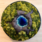 Moosbild "Vulkansee" in einem schwarzen, runden Rahmen. Hier sieht man einen Vulkan aus Modelliermasse (3D) mit einem Vulkansee aus Resin, einen Krater aus schwarzem Sand und grünes, konserviertes Moos. Naturkunst von Moos und Meer in Oberhausen.