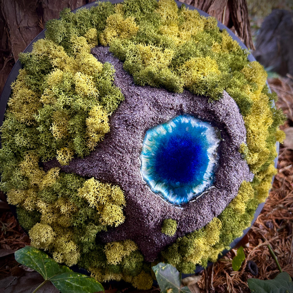 Moosbild "Vulkansee" in einem schwarzen, runden Rahmen. aufgenommen im Wald. Hier sieht man einen Vulkan aus Modelliermasse (3D) mit einem Vulkansee aus Resin, einen Krater aus schwarzem Sand und grünes, konserviertes Moos. Naturkunst von Moos und Meer in Oberhausen.
