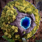 Moosbild "Vulkansee" in einem schwarzen, runden Rahmen. aufgenommen im Wald. Hier sieht man einen Vulkan aus Modelliermasse (3D) mit einem Vulkansee aus Resin, einen Krater aus schwarzem Sand und grünes, konserviertes Moos. Naturkunst von Moos und Meer in Oberhausen.