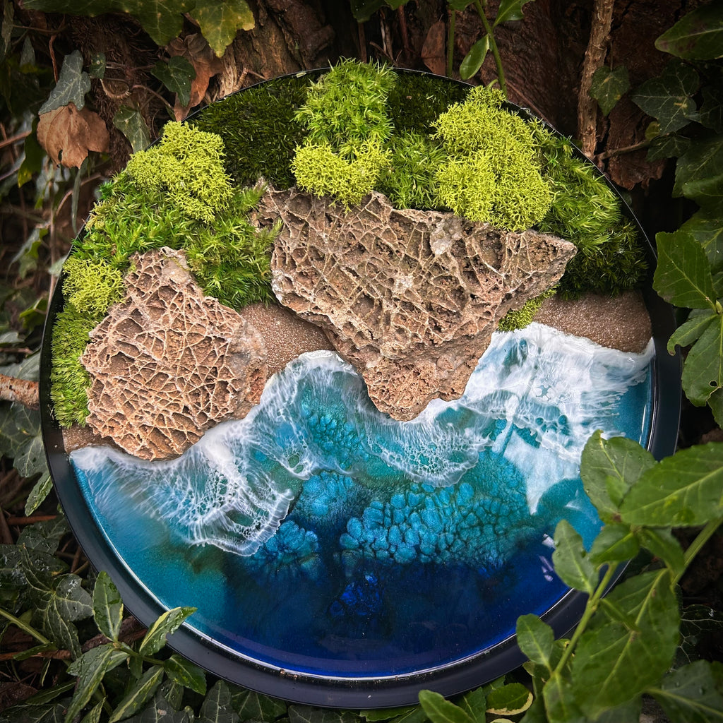 Moosbild "Wellen und Zeit" in einem schwarzen, runden Rahmen. Hier sieht man ein Meer mit Wellen aus Resin, einen Strand mit hellem Sand, Natursteine und grünes, konserviertes Moos. Naturkunst von Moos und Meer in Oberhausen.