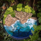 Moosbild "Wellen und Zeit" in einem schwarzen, runden Rahmen. Hier sieht man ein Meer mit Wellen aus Resin, einen Strand mit hellem Sand, Natursteine und grünes, konserviertes Moos. Naturkunst von Moos und Meer in Oberhausen.