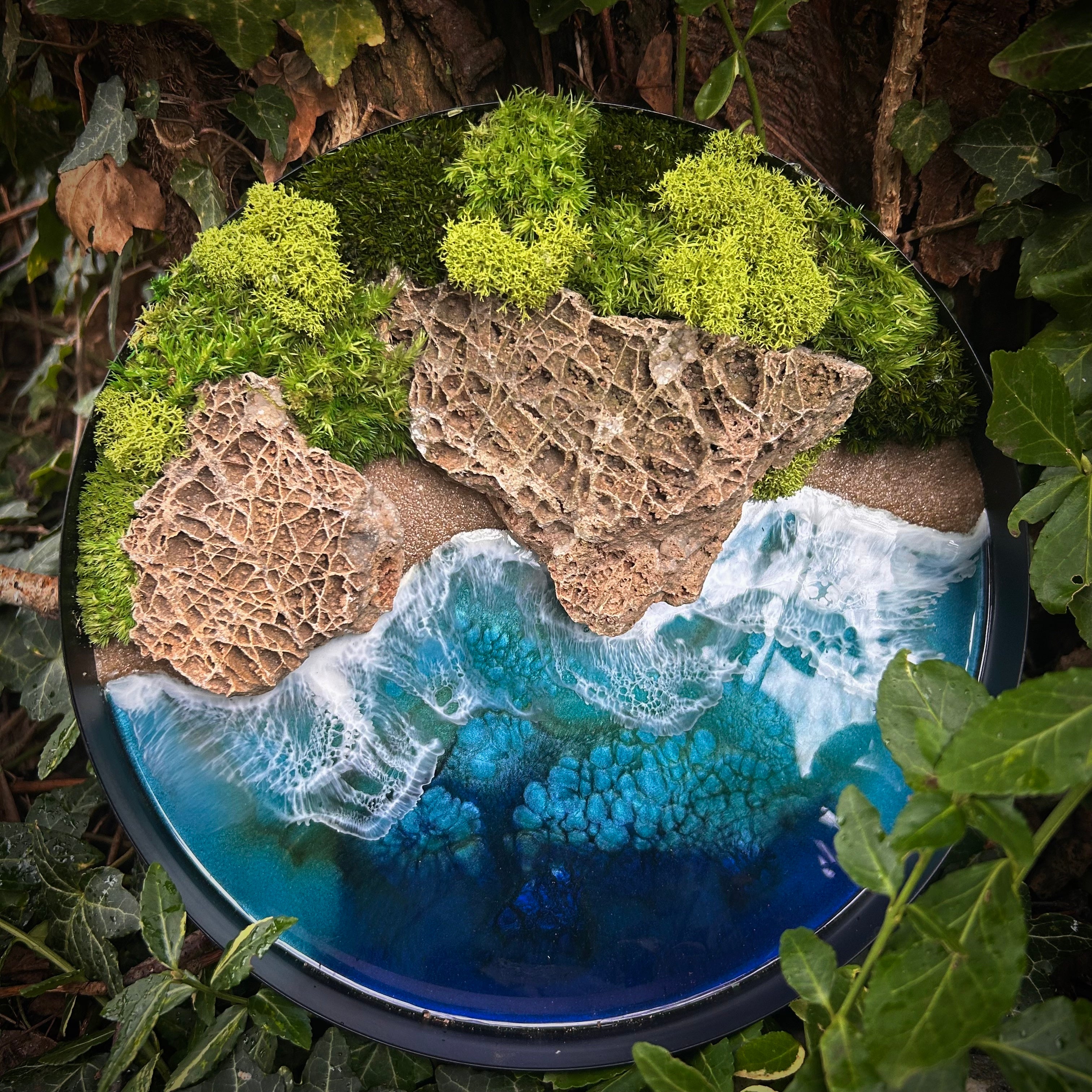 Moosbild "Wellen und Zeit" in einem schwarzen, runden Rahmen. Hier sieht man ein Meer mit Wellen aus Resin, einen Strand mit hellem Sand, Natursteine und grünes, konserviertes Moos. Naturkunst von Moos und Meer in Oberhausen.