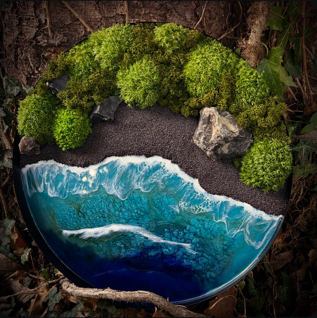 Moosbild "Wildes Paradies" in einem schwarzen, runden Metallrahmen, aufgenommen im Wald. Hier sieht man ein Meer mit Wellen aus Resin, einen Strand mit schwarzem Sand, Natursteine und grünes, konserviertes Moos. Naturkunst von Moos und Meer in Oberhausen.