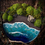 Moosbild "Wildes Paradies" in einem schwarzen, runden Metallrahmen, aufgenommen im Wald. Hier sieht man ein Meer mit Wellen aus Resin, einen Strand mit schwarzem Sand, Natursteine und grünes, konserviertes Moos. Naturkunst von Moos und Meer in Oberhausen.