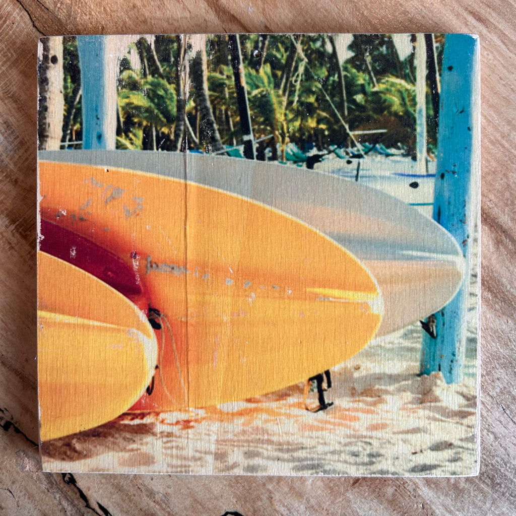 Gelb-orange-farbene Surfbretter liegen auf dem Sand, im Hintergrund sind Palmen. Format 15x15cm. Handgefertigter Fotodruck auf Holz 