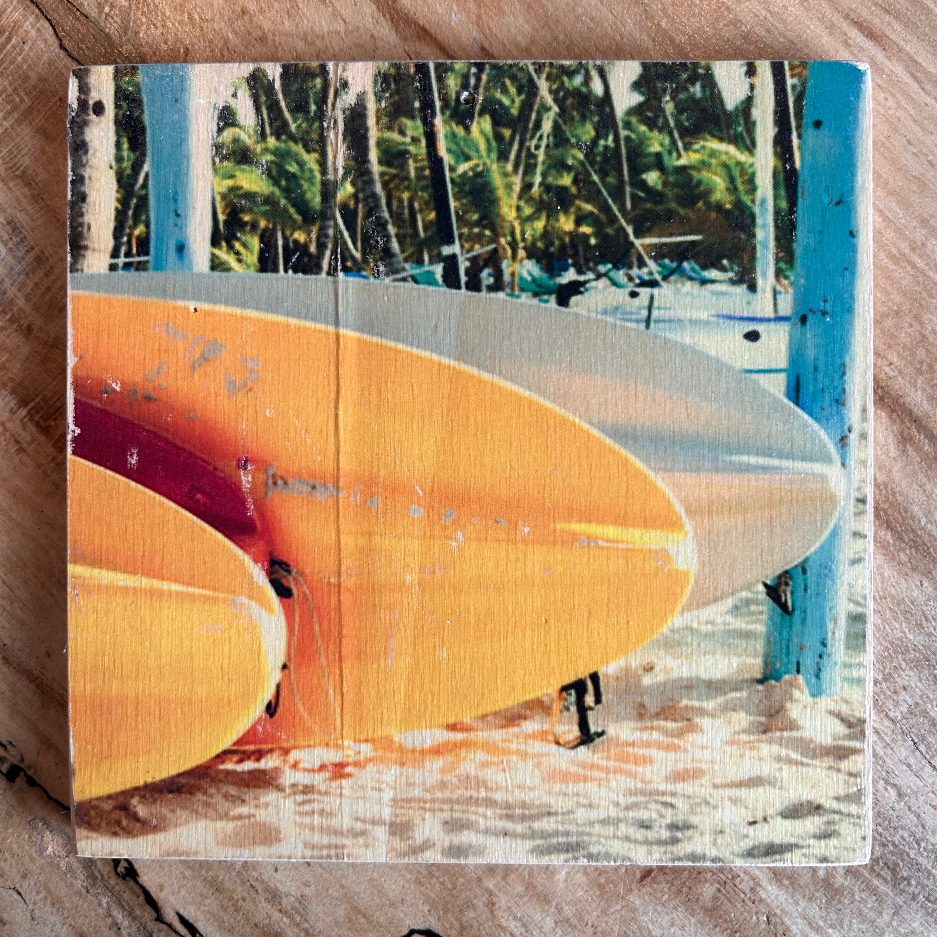 Gelb-orange-farbene Surfbretter liegen auf dem Sand, im Hintergrund sind Palmen. Format 15x15cm. Handgefertigter Fotodruck auf Holz 