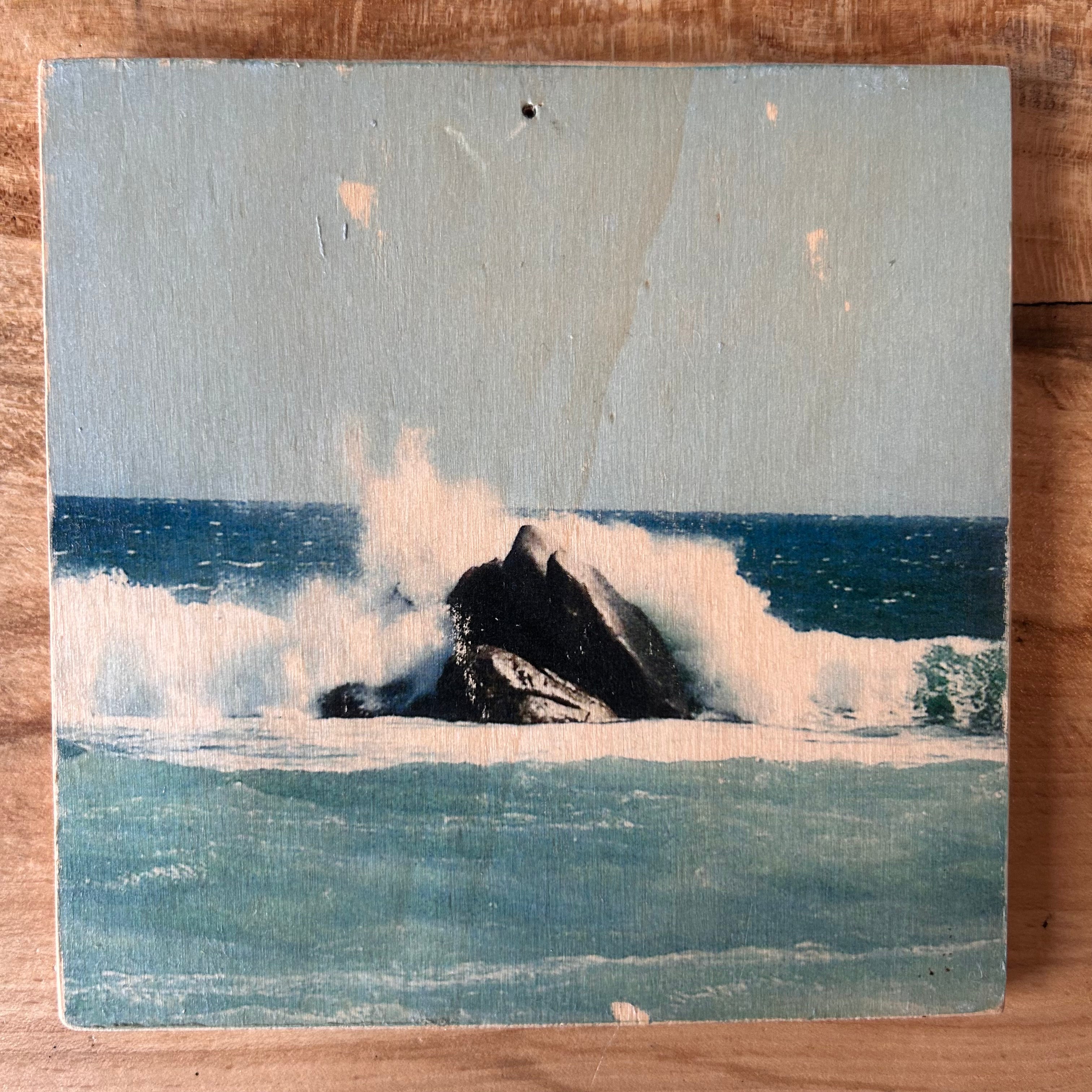Wellen treffen mitten in einem türkis-blauen Ozean auf einen schraz-braunen Felsen und die Gischt der Wellen spritzen. Format 15x15cm. Handgefertigter Fotodruck auf Holz 