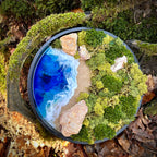 Moosbild "Weitblick" in einem schwarzen, runden Rahmen, aufgenommen im Wald. Hier sieht man ein Meer mit Wellen aus Resin, einen Strand mit hellem Sand, Natursteine und grünes, konserviertes Moos. Naturkunst von Moos und Meer in Oberhausen.