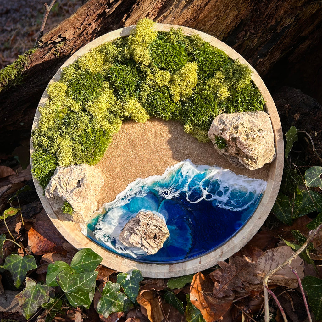 Rundes Naturkunstwerk mit Moos, Sand, Steinen und blau-türkisem Resinmeer, das eine ruhige Küstenlandschaft mit Wellen, Felsen und Wald darstellt.