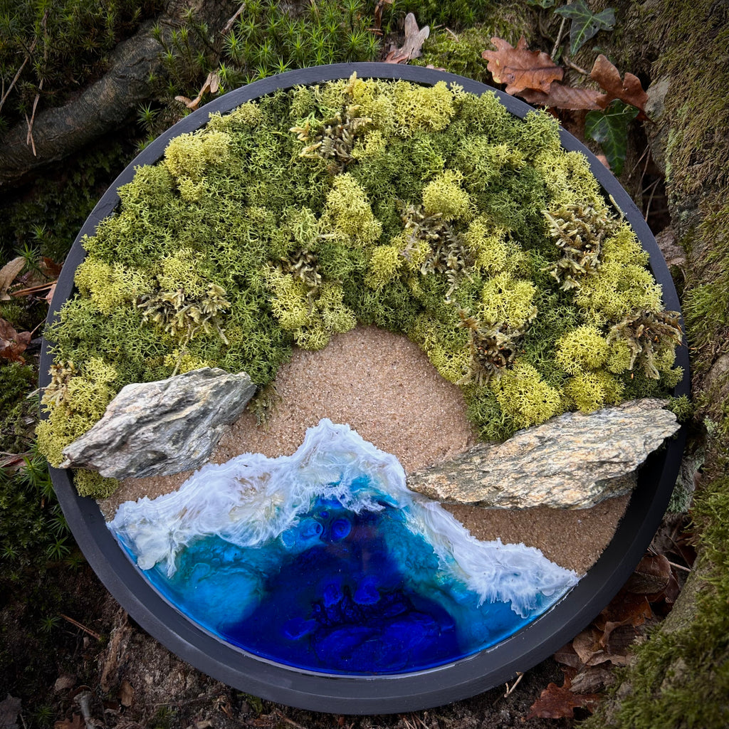 Rundes Moos- und Meerbild mit spitzen, schmalen Felsen, grünem Moos, Sandstrand und tiefblauem Resin-Meer mit sanften Wellen, eingefasst in einem dunklen Rahmen.