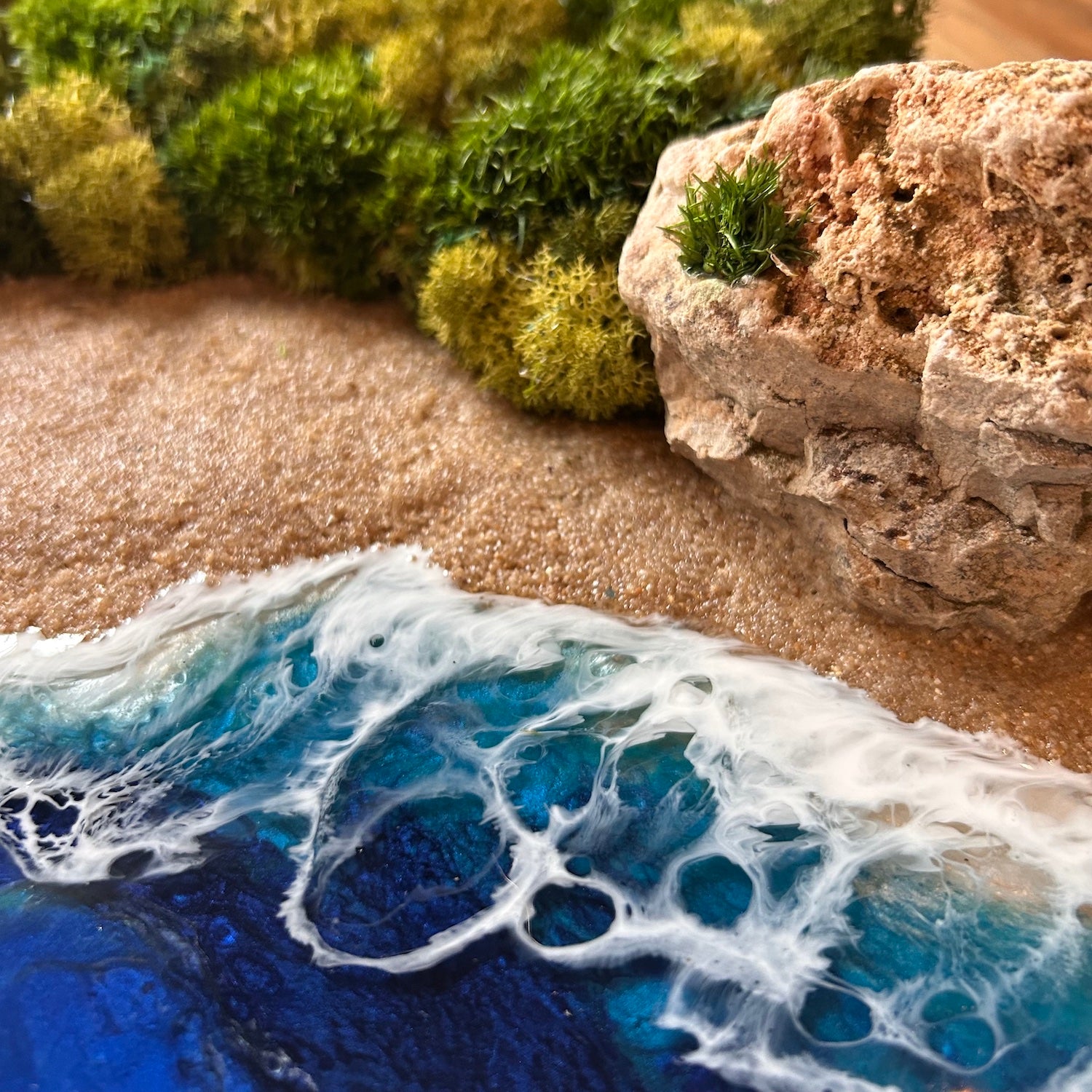 Detailaufnahme des Moosbild "Goldener Strand" . Hier sieht man ein Meer mit Wellen aus Resin, einen Strand mit hellem Sand, Natursteine und grünes, konserviertes Moos. Naturkunst von Moos und Meer in Oberhausen.