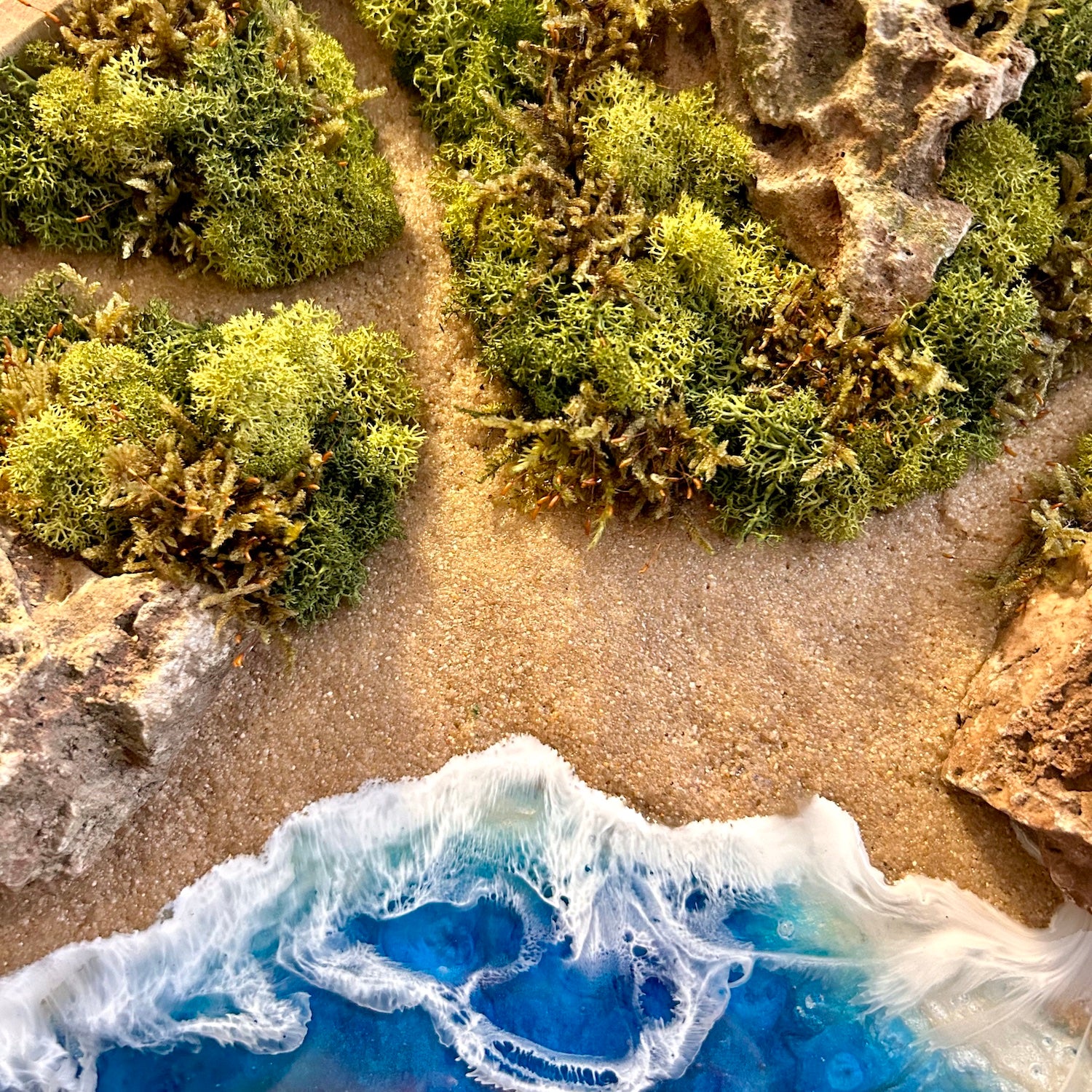 Detailaufnahme des Moosbild "Meerpfade" . Hier sieht man ein Meer mit Wellen aus Resin, einen Strand mit hellem Sand, kleine Pfade zwischen den Moosbereichen, Natursteine und grünes, konserviertes Moos. Naturkunst von Moos und Meer in Oberhausen.