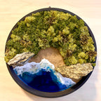 Moosbild "Strandwächter" in einem schwarzen, runden Rahmen. Hier sieht man ein Meer mit Wellen aus Resin, einen Strand mit hellem Sand, Natursteine und grünes, konserviertes Moos. Naturkunst von Moos und Meer in Oberhausen.