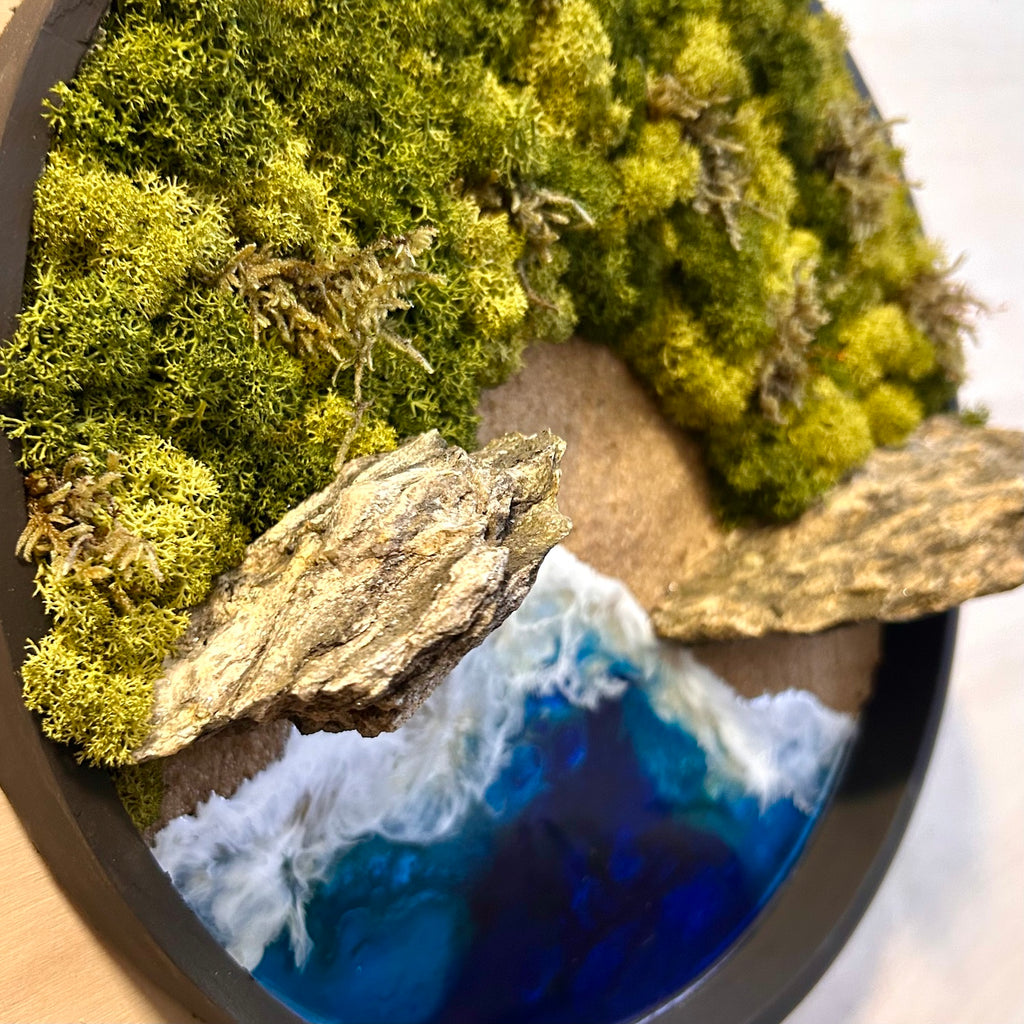 Seitliche Aufnahme des Moosbild "Strandwächter" in einem schwarzen, runden Rahmen. Hier sieht man ein Meer mit Wellen aus Resin, einen Strand mit hellem Sand, Natursteine und grünes, konserviertes Moos. Naturkunst von Moos und Meer in Oberhausen.