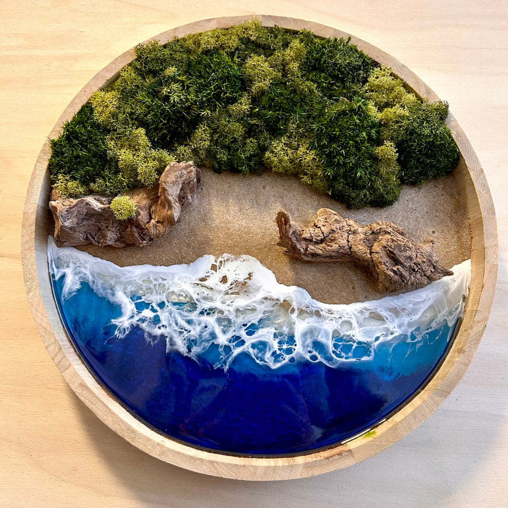 Moosbild "Treibholzstrand" in einem runden Rahmen aus Bambus. Hier sieht man ein Meer mit Wellen aus Resin, einen Strand mit hellem Sand, Treibholz und grünes, konserviertes Moos. Naturkunst von Moos und Meer in Oberhausen.
