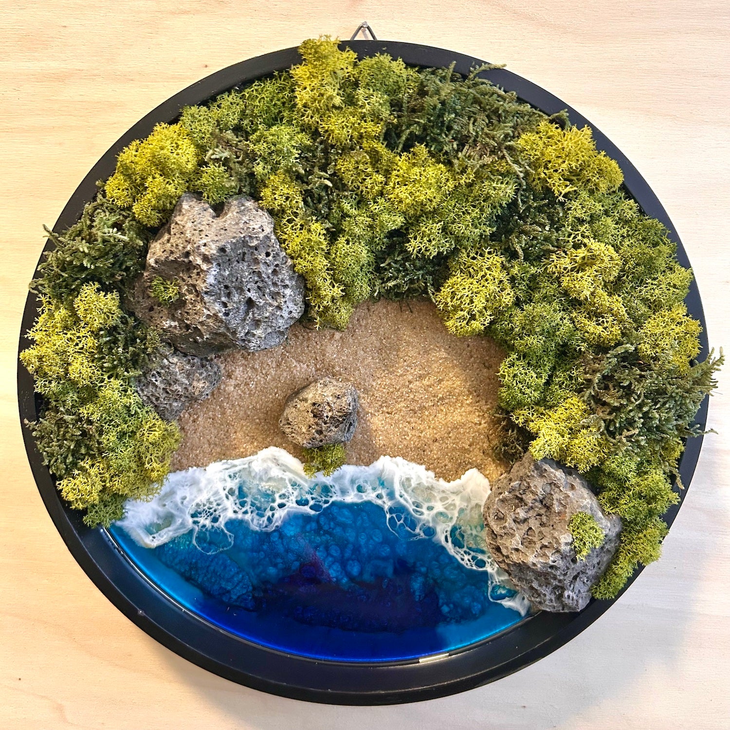 Moosbild "Versteckte Bucht" in einem runden, schwarzen Rahmen. Hier sieht man ein Meer mit Wellen aus Resin, einen Strand mit hellem Sand, Natursteine und grünes, konserviertes Moos. Naturkunst von Moos und Meer in Oberhausen.