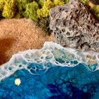 Detailaufnahme des Moosbild "Versteckte Bucht" . Hier sieht man ein Meer mit Wellen aus Resin, einen Strand mit hellem Sand, Natursteine und grünes, konserviertes Moos. Naturkunst von Moos und Meer in Oberhausen.