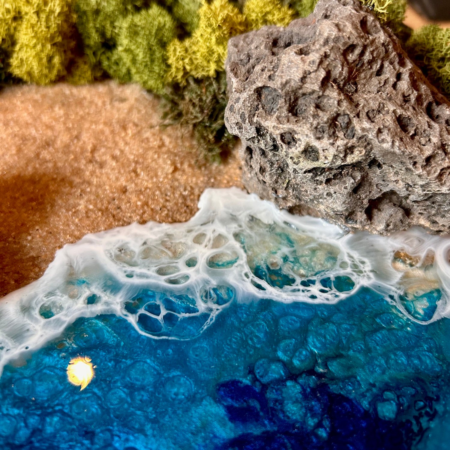 Detailaufnahme des Moosbild "Versteckte Bucht" . Hier sieht man ein Meer mit Wellen aus Resin, einen Strand mit hellem Sand, Natursteine und grünes, konserviertes Moos. Naturkunst von Moos und Meer in Oberhausen.