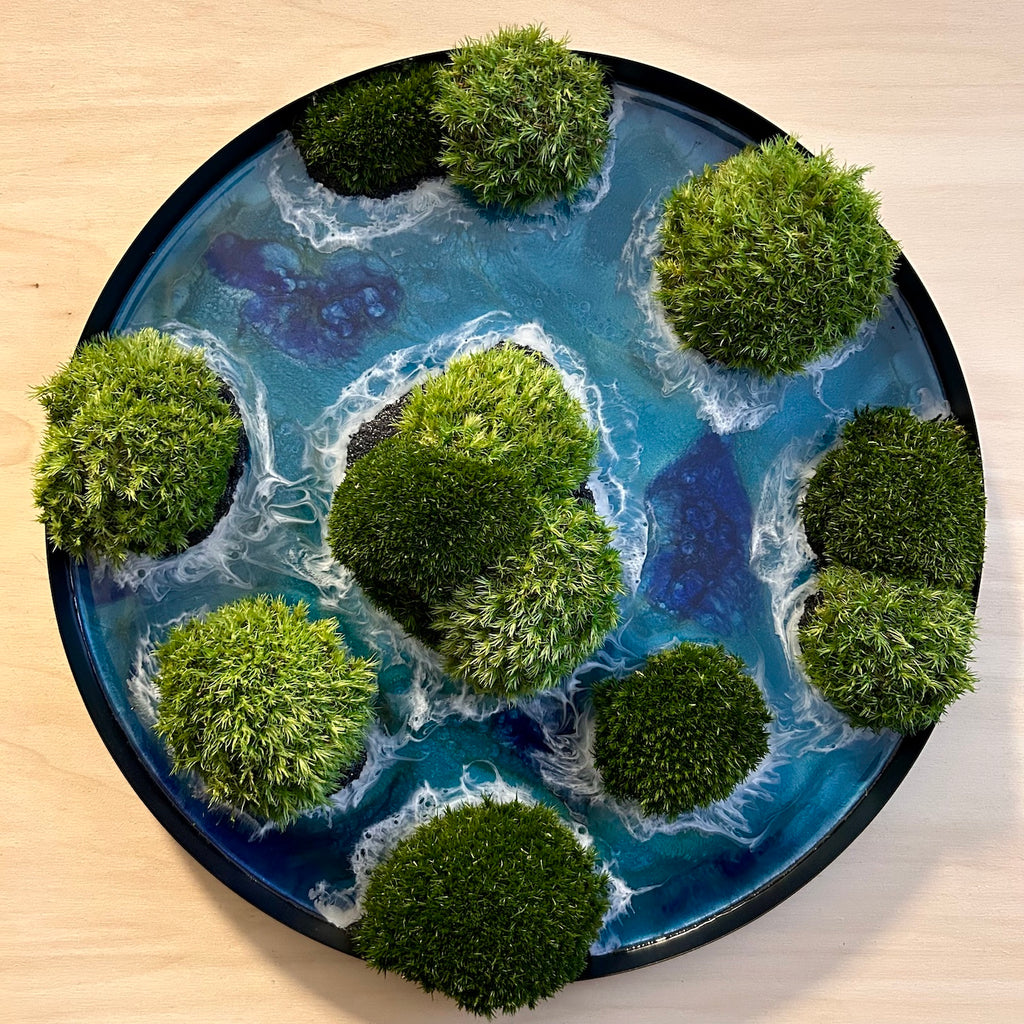 Moosbild "Waterworld" ine inem runden Rahmen aus schwarzem Metall. Hier sieht man ein Meer mit Wellen aus Resin, Inseln mit schwarzem Sand und grünem, konserviertem Moos. Naturkunst von Moos und Meer in Oberhausen.