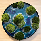 Moosbild "Waterworld" ine inem runden Rahmen aus schwarzem Metall. Hier sieht man ein Meer mit Wellen aus Resin, Inseln mit schwarzem Sand und grünem, konserviertem Moos. Naturkunst von Moos und Meer in Oberhausen.