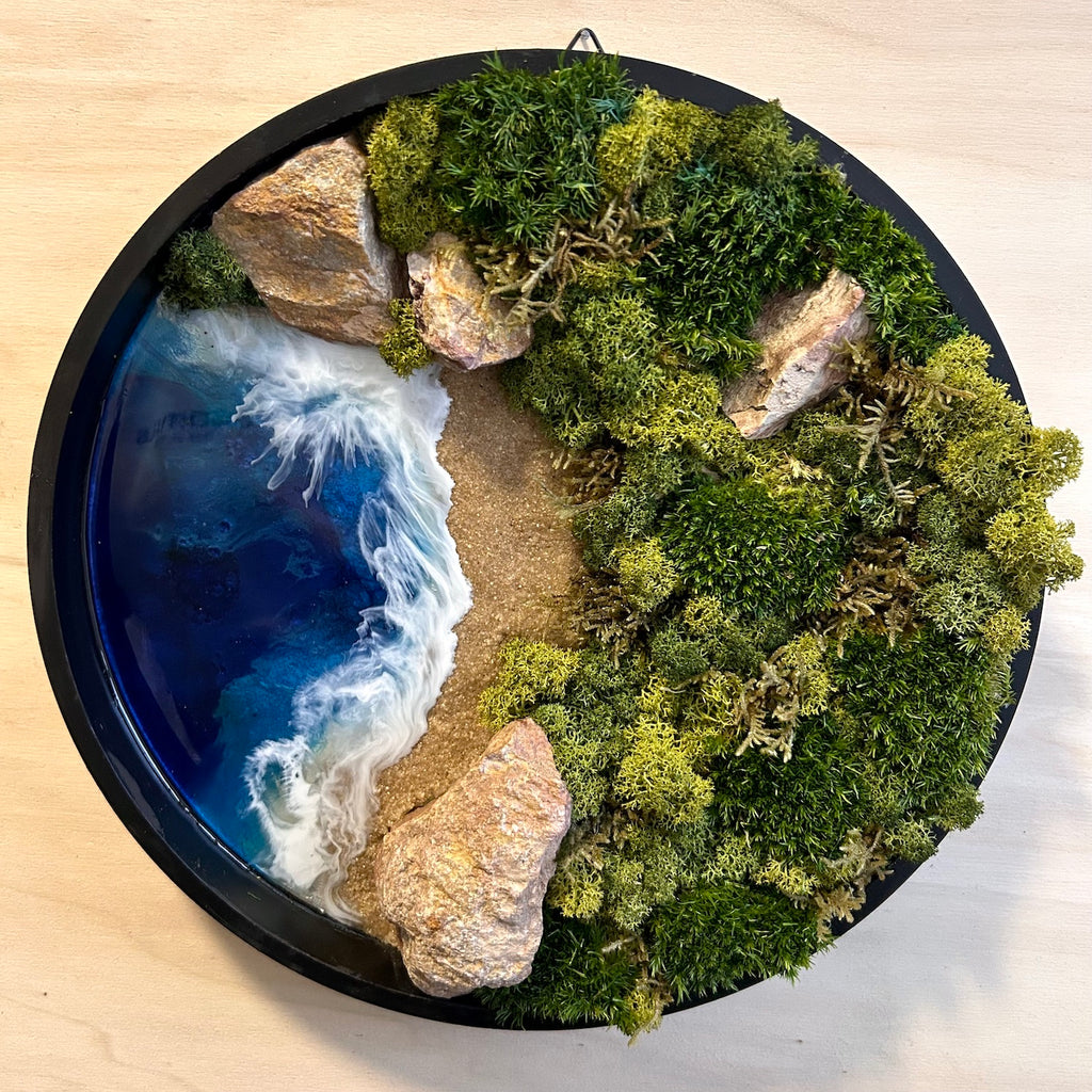 Moosbild "Weitblick" in einem runden, schwarzen Rahmen. Hier sieht man ein Meer mit Wellen aus Resin, einen Strand mit hellem Sand, Natursteine und grünes, konserviertes Moos. Naturkunst von Moos und Meer in Oberhausen.