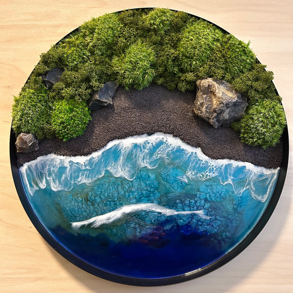 Moosbild "Wildes Paradies" in einem schwarzen, runden Metallrahmen. Hier sieht man ein Meer mit Wellen aus Resin, einen Strand mit schwarzem Sand, Natursteine und grünes, konserviertes Moos. Naturkunst von Moos und Meer in Oberhausen.