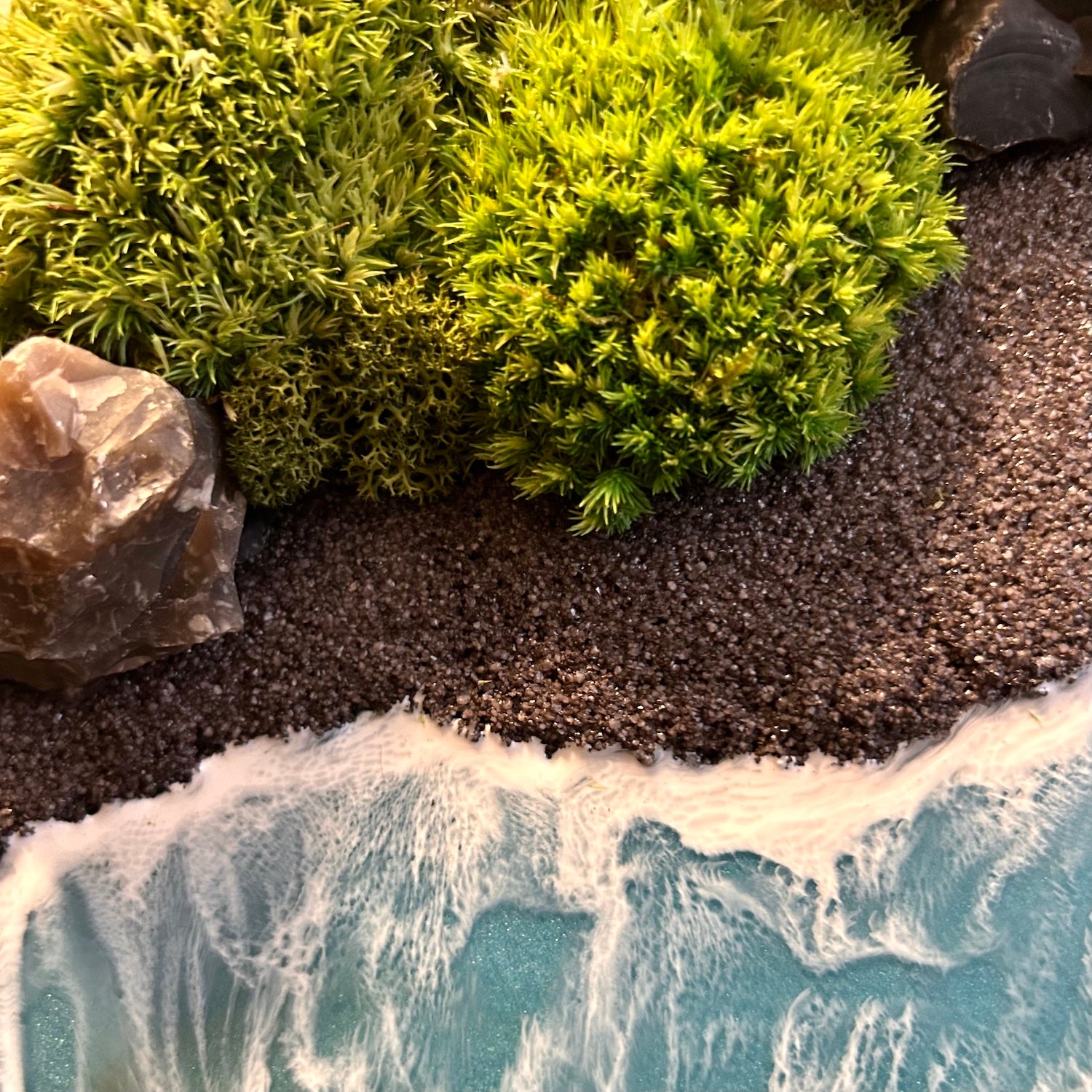 Detailaufnahme des Moosbild "Wildes Paradies". Hier sieht man ein Meer mit Wellen aus Resin, einen Strand mit schwarzem Sand, Natursteine und grünes, konserviertes Moos. Naturkunst von Moos und Meer in Oberhausen.