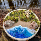 Rundes Moos- und Meerbild mit grünen Mooslandschaften, sandfarbenen Wegen, Natursteinen und tiefblauem Resin-Meer mit schäumenden Wellen, eingebettet in einen Holzrahmen. Hier aus einer leicht schrägen Perspektive fotografiert. 