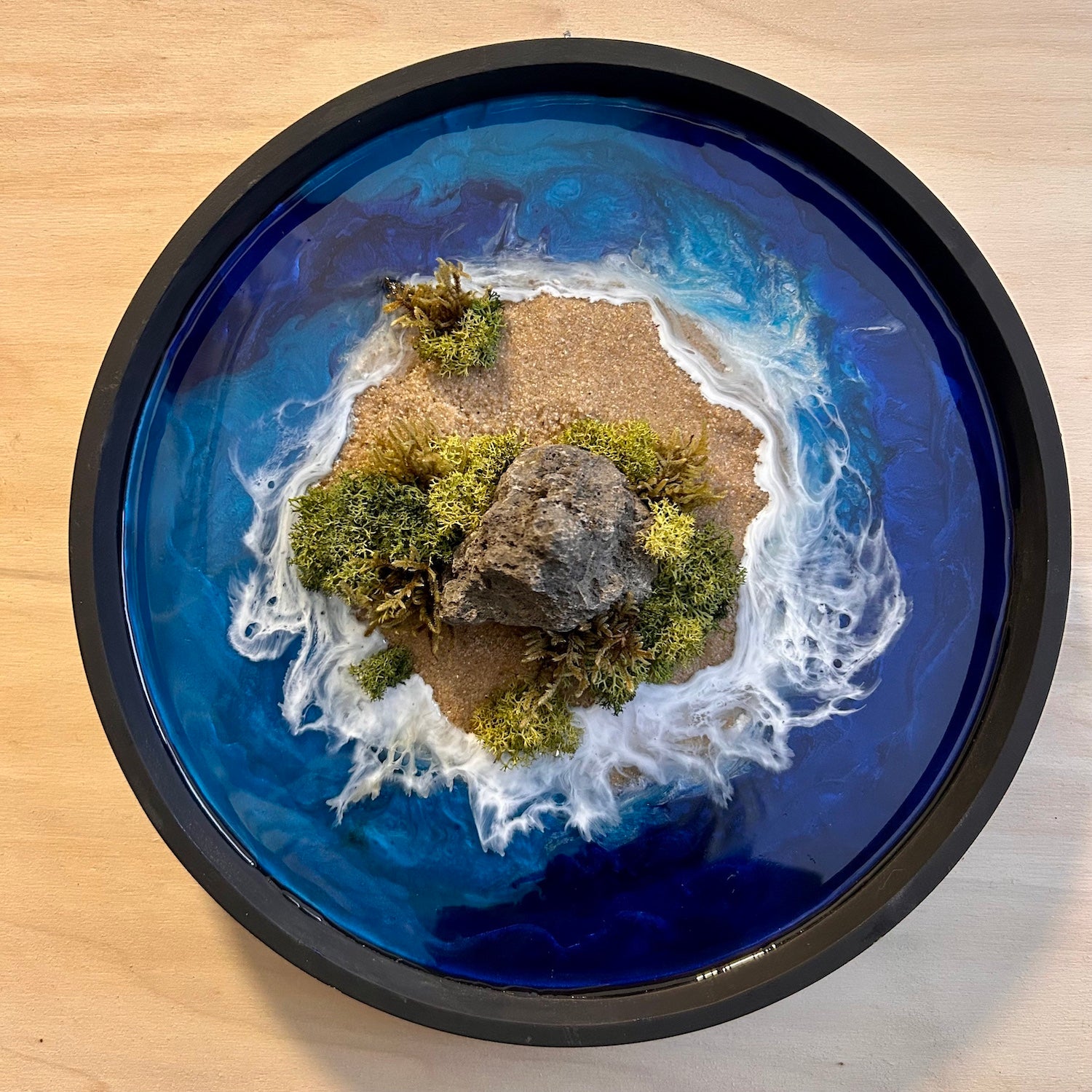 Moosbild "Einsame Insel" in einem schwarzen, runden Rahmen. Hier sieht man ein Meer mit Wellen aus Resin, einen Strand mit hellem Sand, Natursteine und grünes, konserviertes Moos. Naturkunst von Moos und Meer in Oberhausen.