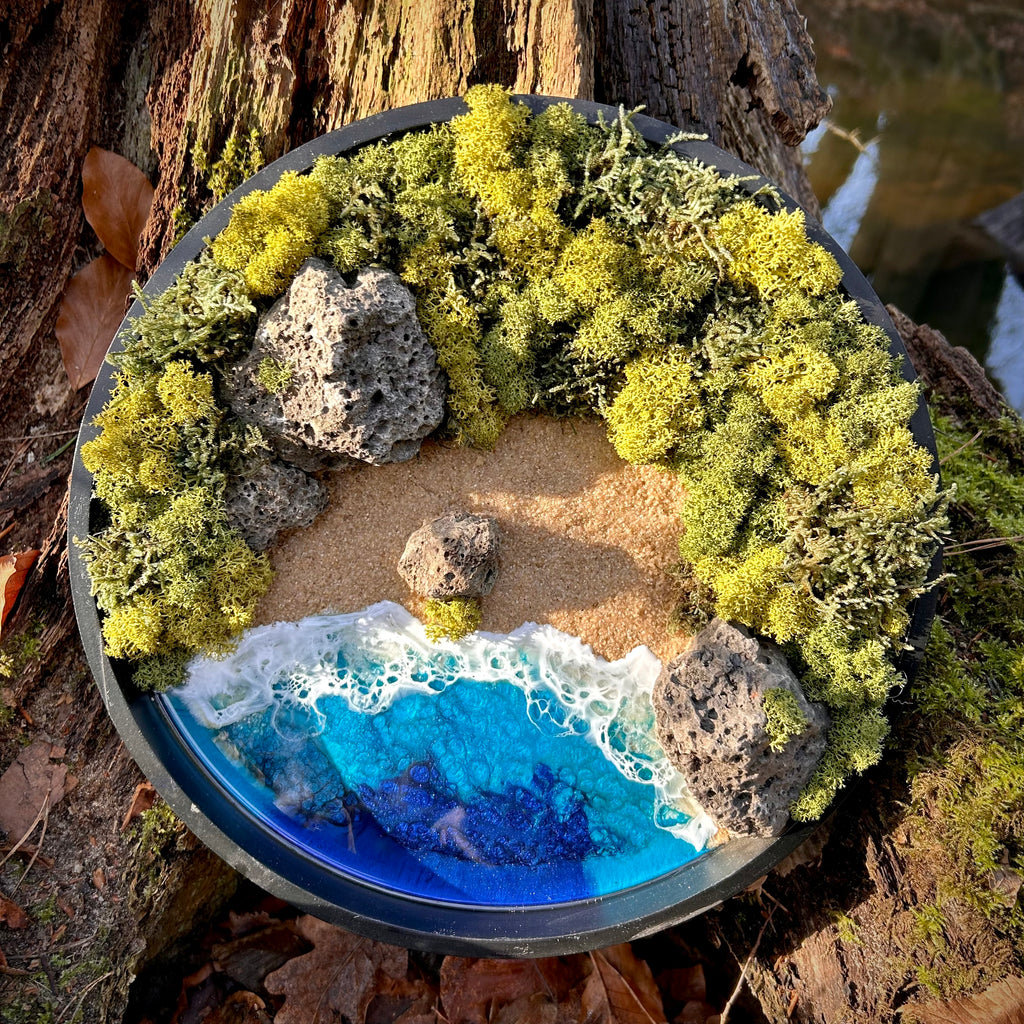 Moosbild "Versteckte Bucht" in einem schwarzen, runden Rahmen, aufgenommen im Wald. Hier sieht man ein Meer mit Wellen aus Resin, einen Strand mit hellem Sand, Natursteine und grünes, konserviertes Moos. Naturkunst von Moos und Meer in Oberhausen.